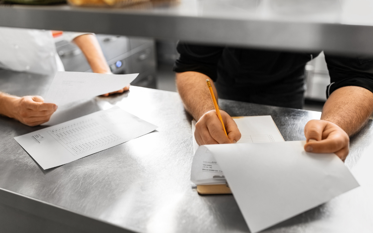 two peoples hands are seen looking at paperwork and writing on a clip board in a restaurant kitchen serving area