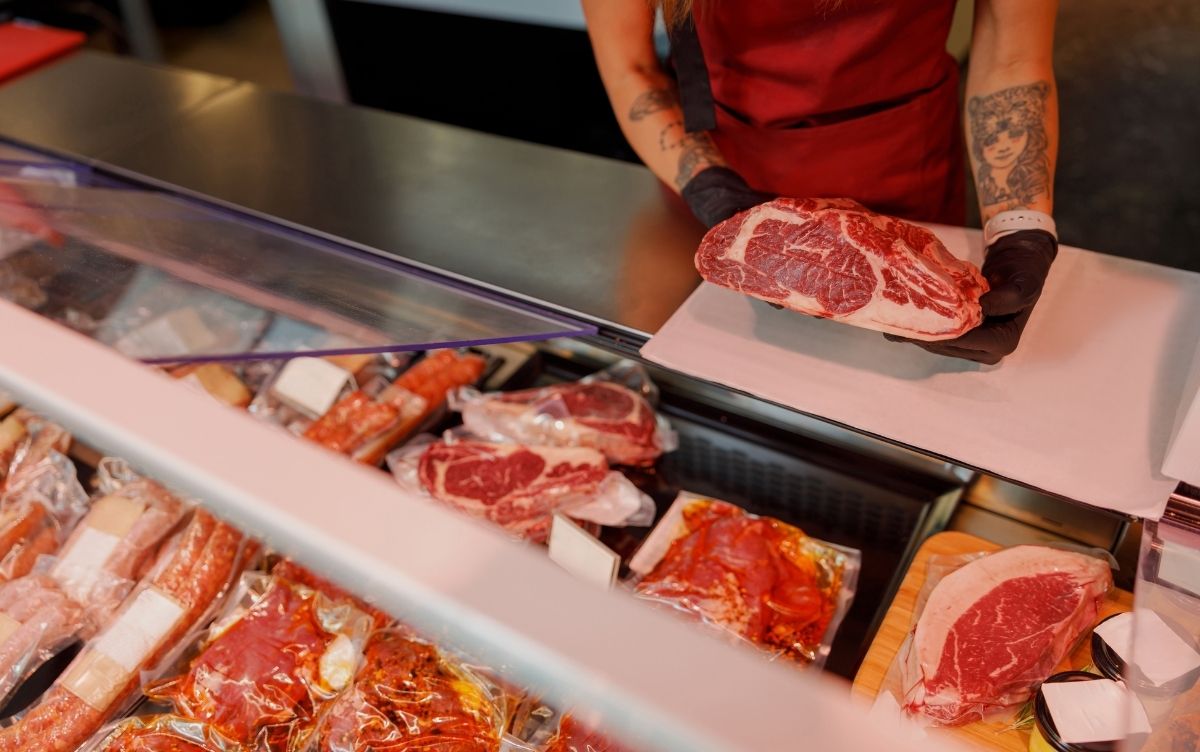 Butcher wearing gloves and apron holding a large marbled cut of beef behind a meat counter displaying various packaged and tray-laid cuts.