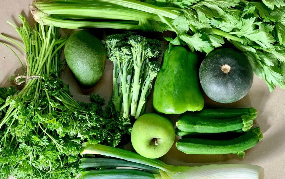 A variety of fresh green vegetables and fruits, including celery, avocado, broccoli, bell pepper, zucchini, and green apples, neatly arranged on a light surface