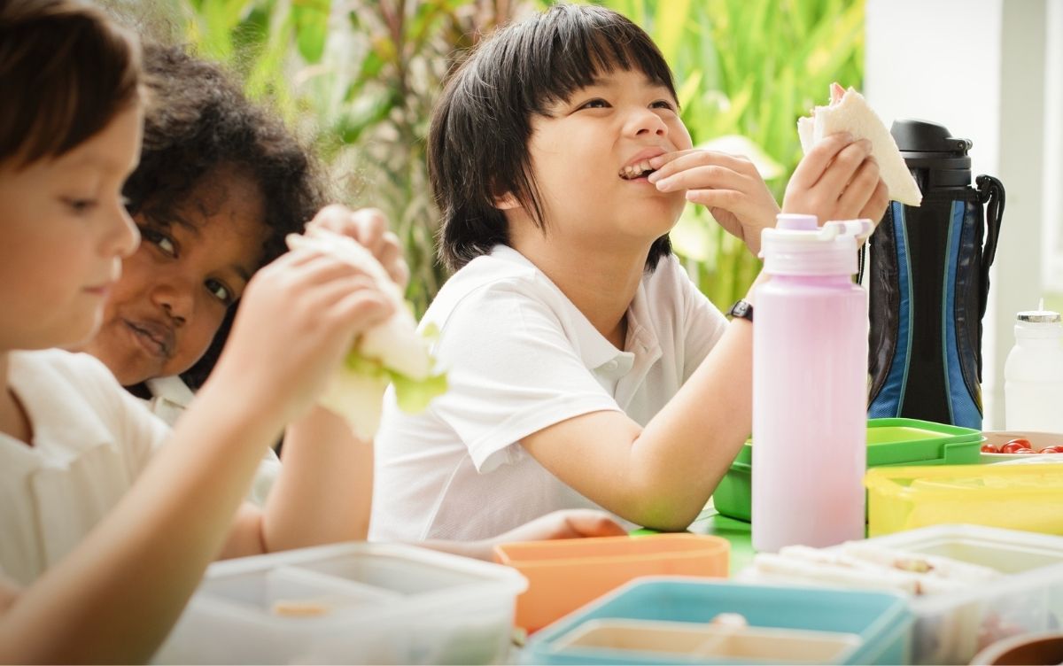 Three children sitting eating a packed lunch of sandwiches.