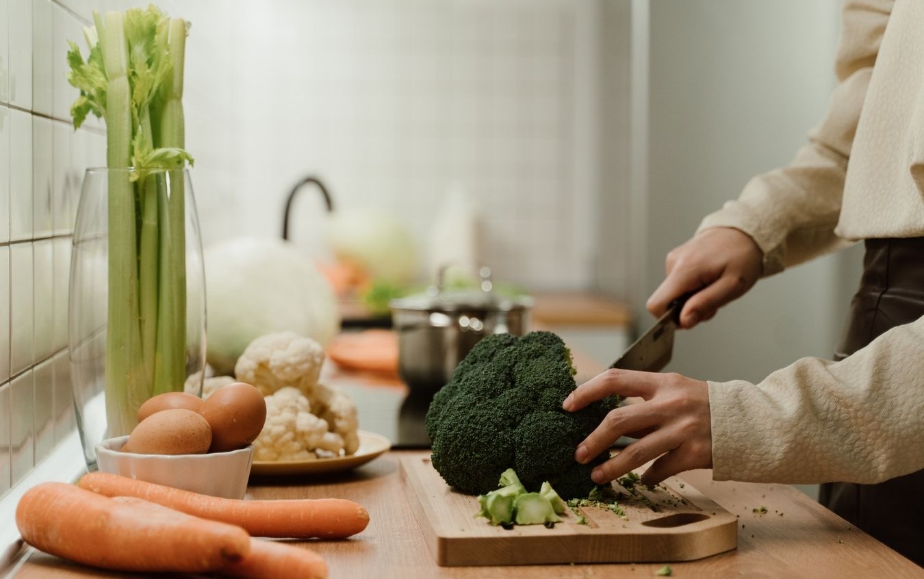 Person chopping broccoli on a wooden cutting board surrounded by carrots, eggs, cauliflower, and celery, with kitchen items and a pot on the stove in the background.