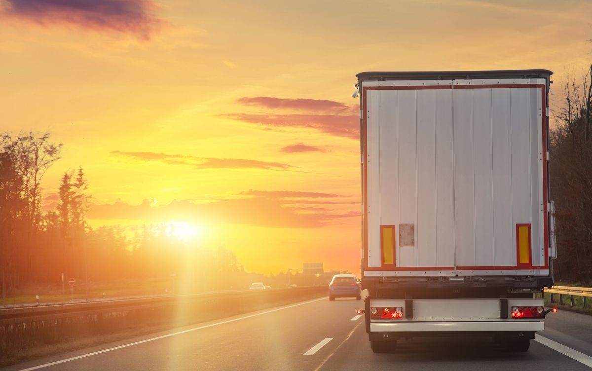 Rear view of a large delivery truck driving on a highway during sunset, with warm orange and yellow hues in the sky and other vehicles visible ahead.