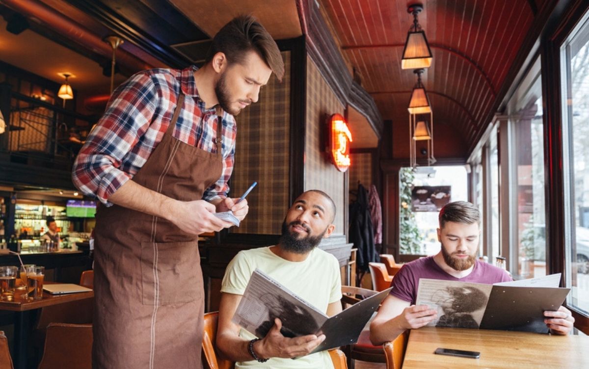 Two men sitting at a table in a restaurant looking at menus. One is speaking to the waiter.