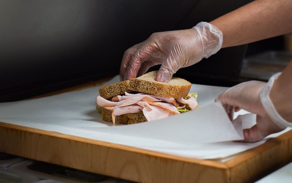 Hands in gloves preparing a sandwich with turkey and lettuce on whole grain bread on a kitchen counter.