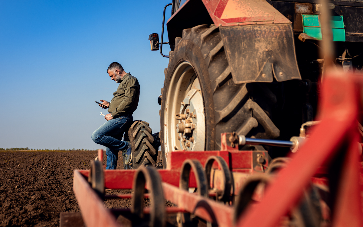 Male farmer checks for news on mobile phone standing next to tractor wheel on a ploughed field, with blue sky above