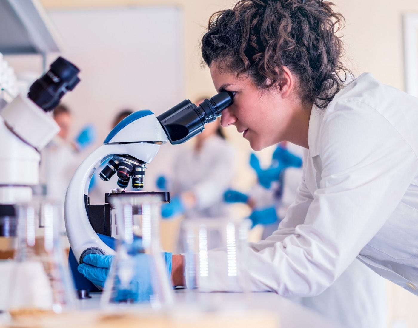 Person in a lab coat looking through a microscope in a busy laboratory, with other lab-coated individuals and equipment like beakers and microscopes in the background.