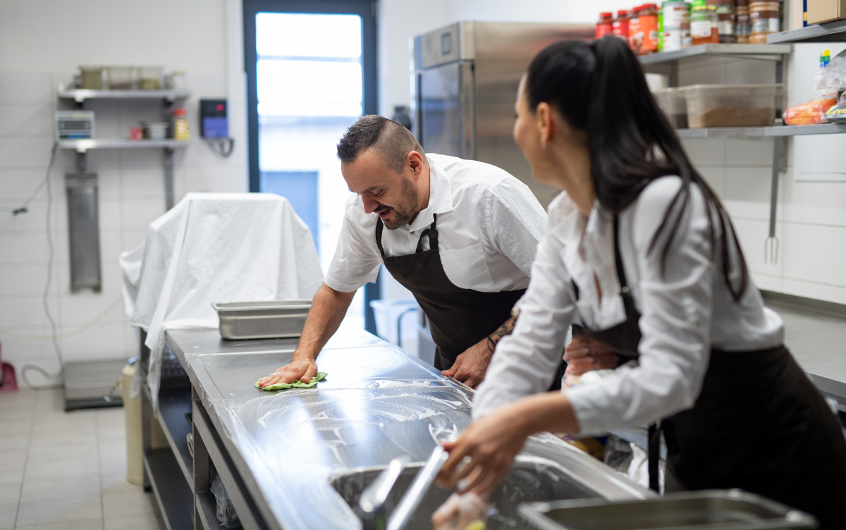 A make and female member of kitchen staff are cleaning food preparation areas with soap and a cloth