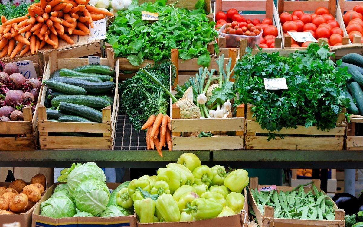 A market stall displays a colourful assortment of fresh vegetables in wooden crates, including leafy greens, tomatoes, cucumbers, carrots, bell peppers, and green beans, with handwritten labels marking types and prices.