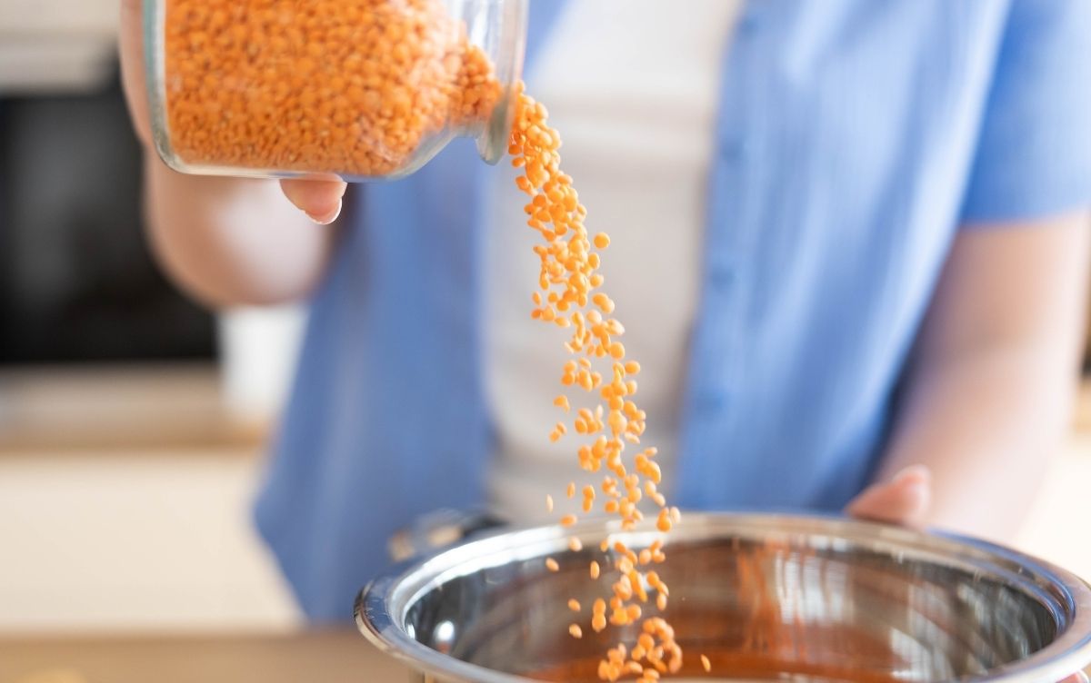 A person pours lentils from a clear container into a stainless steel bowl in a kitchen.