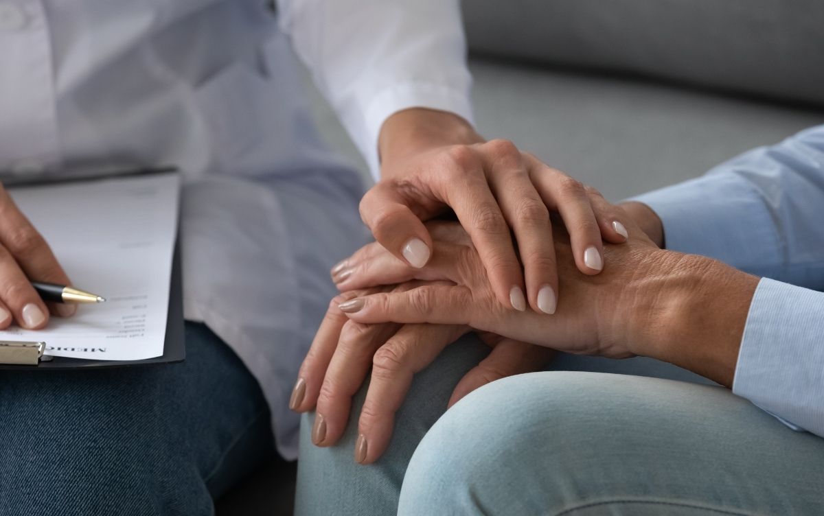 Close-up of two people sitting together, with a medical professional holding a clipboard and gently holding the other person's hands in a supportive gesture.