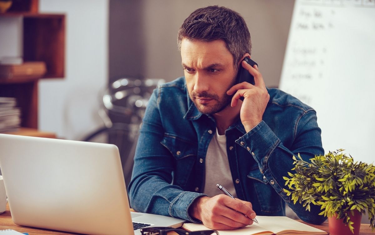 Man sitting while looking at a laptop. He's also on the phone.