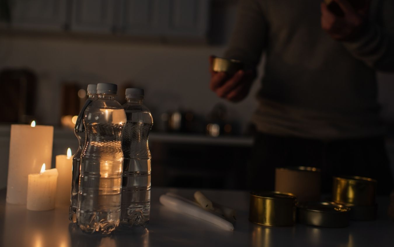 Dimly lit indoor scene with two bottles of water, several lit candles, a person holding a small can, additional cans on the table, and a flashlight or cylindrical object.