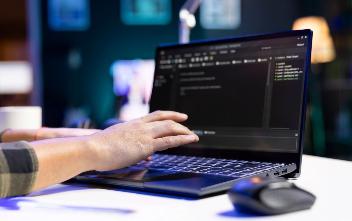 Person typing on a laptop displaying a dark-themed code editor, in a tech-focused indoor workspace with blurred background elements.