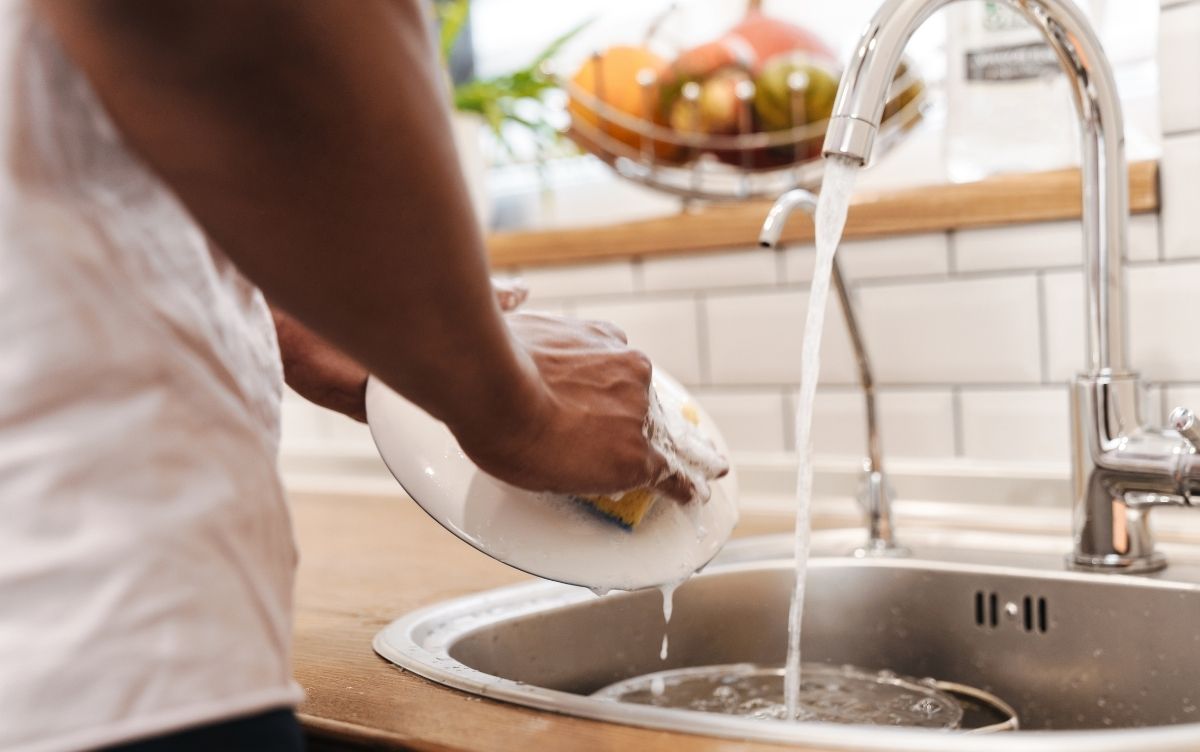 Man washing a plate at a kitchen sink. The tap is on.