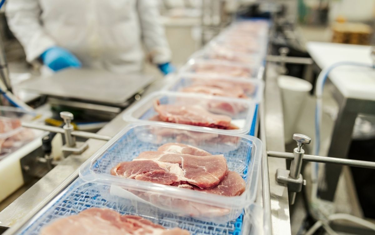 Trays of raw meat on a conveyor belt in a meat processing facility, with a worker in protective clothing and gloves in the background, illustrating industrial food packaging operations