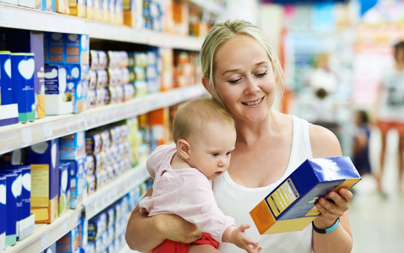 erson holding a baby while shopping in a grocery store aisle, examining a product box with shelves stocked with various items.
