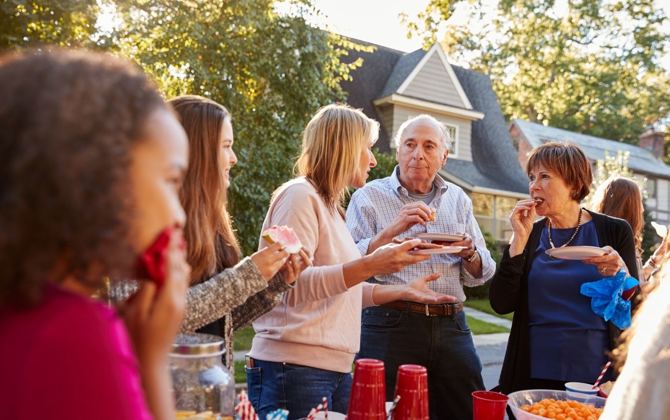 Group of people gathered outdoors around a table with food and red plastic cups, with trees and a house in the background. Faces are blurred for privacy