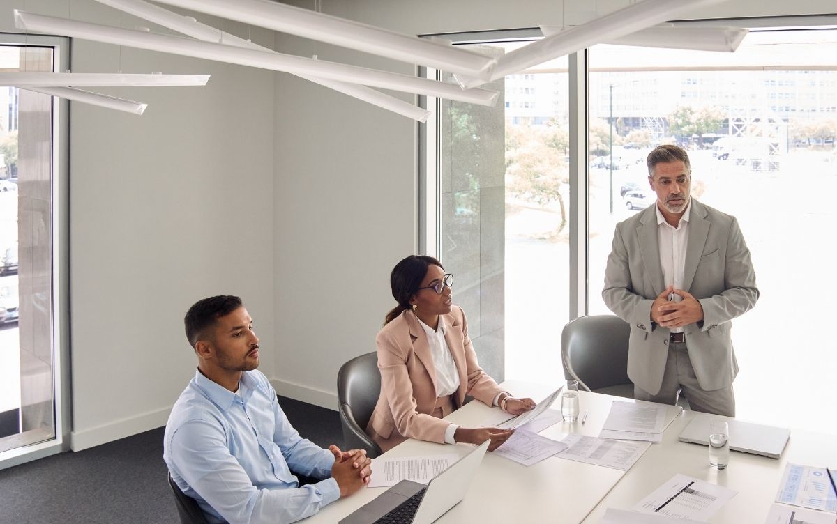 Three office workers in a meeting room, with one person standing and speaking while two others sit at a table with laptops and documents, in a bright, modern workspace.