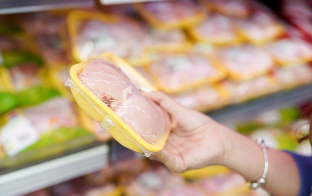 Hand holding a packaged tray of raw chicken in a supermarket, with multiple similar packages displayed in the background.