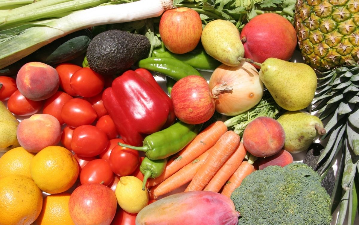 A colorful assortment of fresh fruits and vegetables including tomatoes, carrots, bell peppers, avocados, apples, pears, peaches, oranges, lemons, onions, green chili peppers, broccoli, celery stalks, and a pineapple.