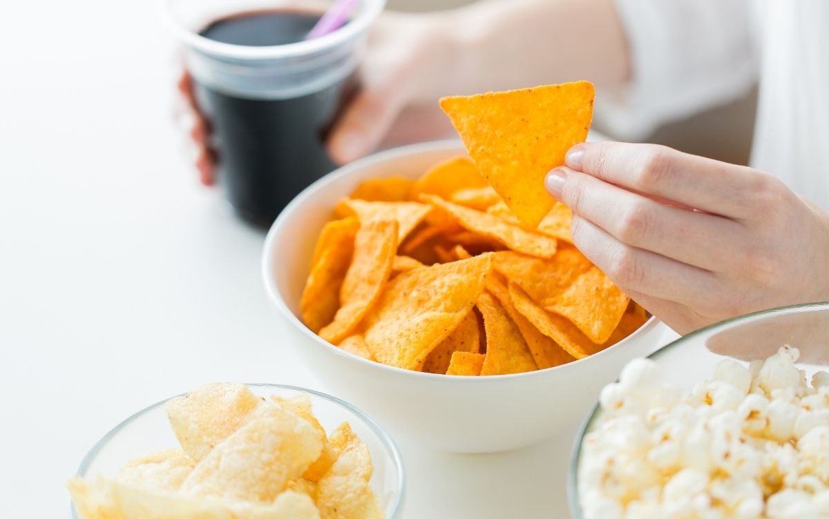 A person holding a tortilla chip, with a bowl of chips, a cup of coffee, and a bowl of popcorn on a white table.