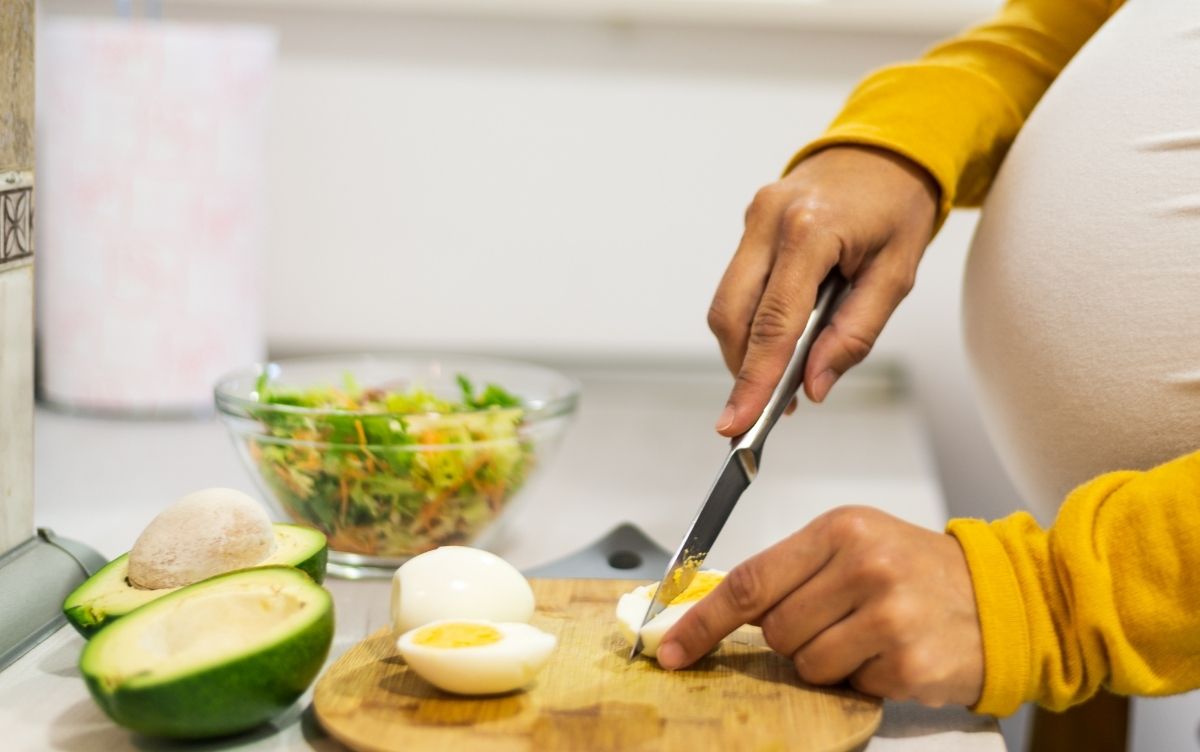 Person preparing a salad, slicing a boiled egg on a wooden cutting board, with avocado and mushrooms nearby.