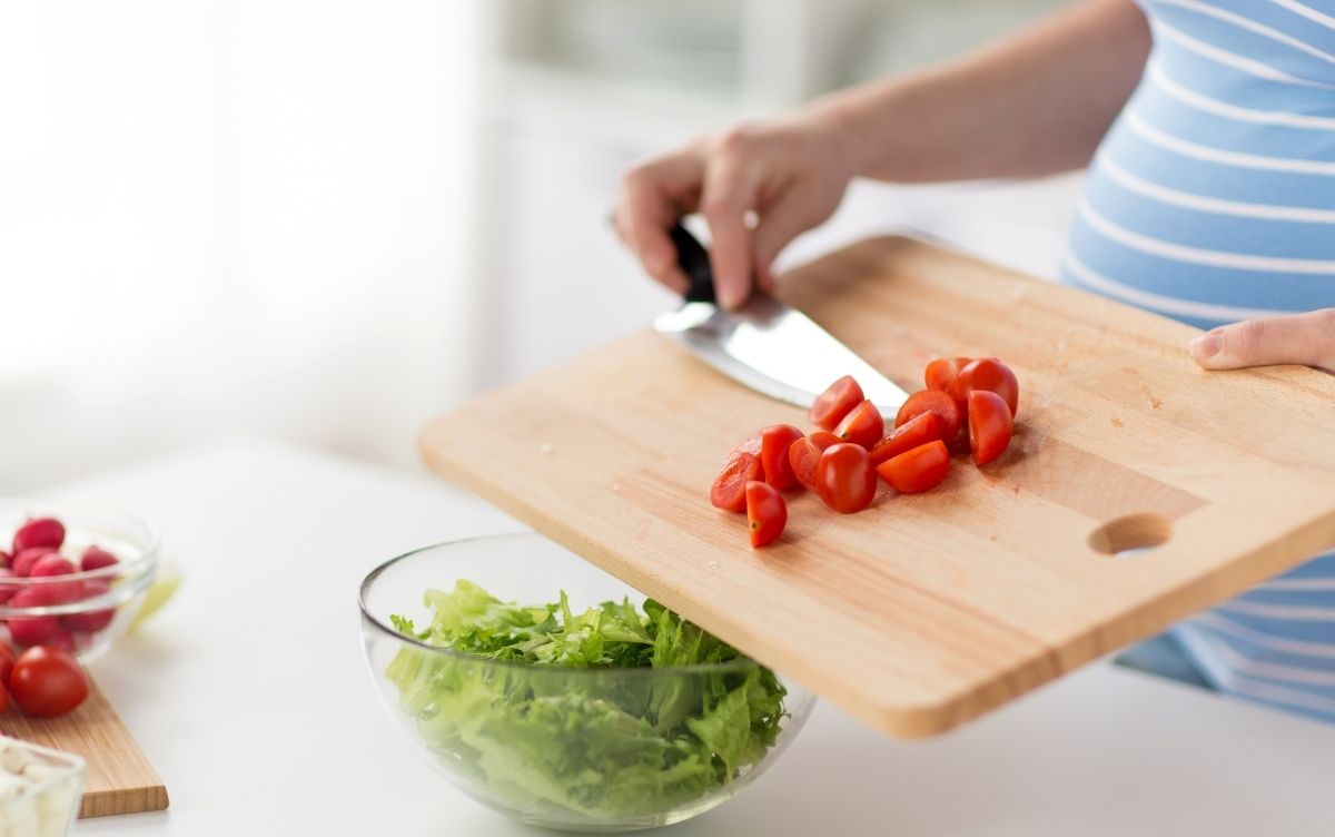 Pregnant woman with a chopping board and adding tomatoes to lettuce.