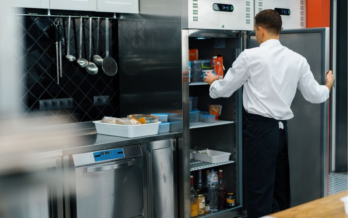 Chef in a professional kitchen wearing a white jacket and black apron, organizing items in a stainless steel refrigerator, with utensils and appliances visible in the background.