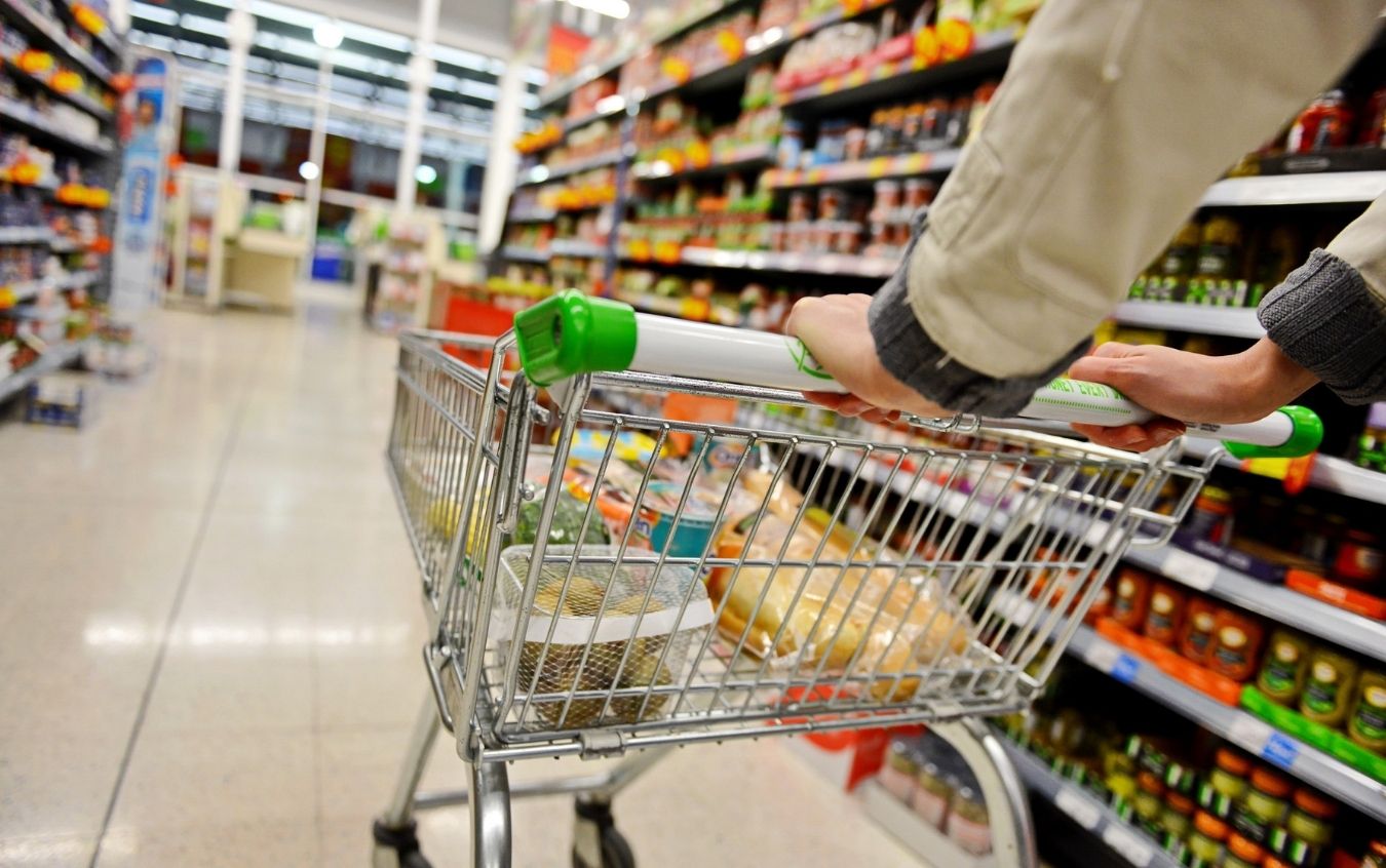 Person pushing a shopping cart down a grocery store aisle, with shelves on both sides stocked with various products.