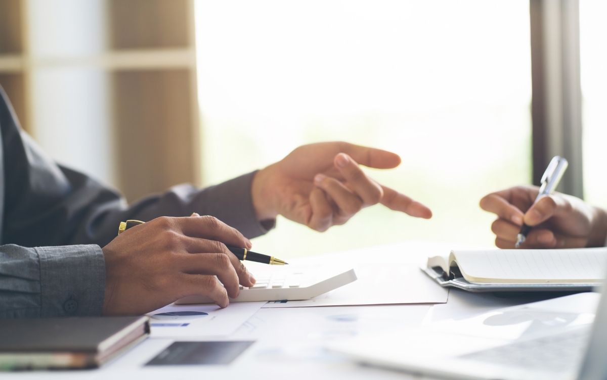 Close up of one man pointing and holding a pen while the other writes in a notebook.