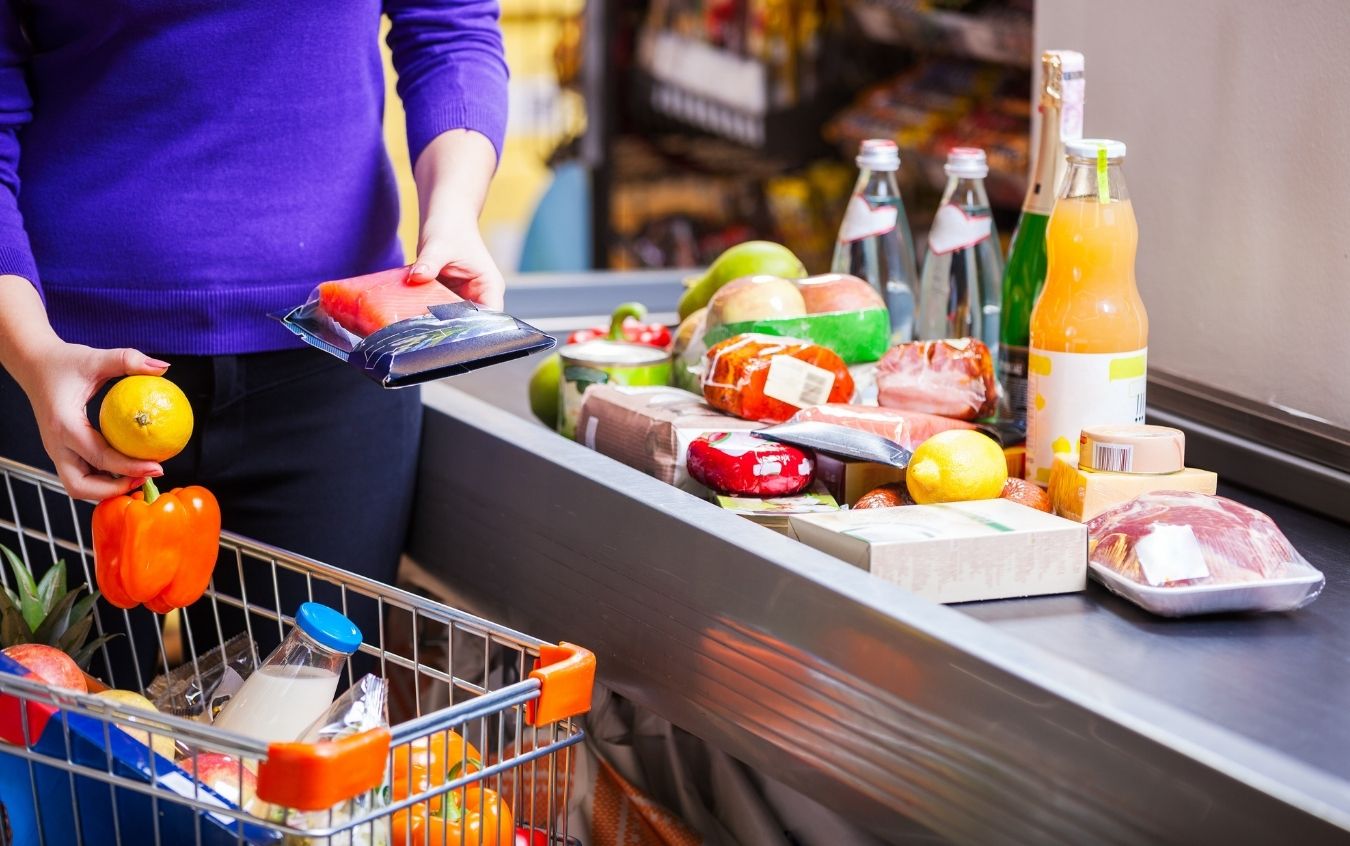 Person in a purple sweater holding a lemon and packaged food at a grocery store checkout. Shopping cart contains a pineapple, orange bell pepper, and other groceries. Conveyor belt displays bottled drinks, packaged meat, fruits, and vegetables.