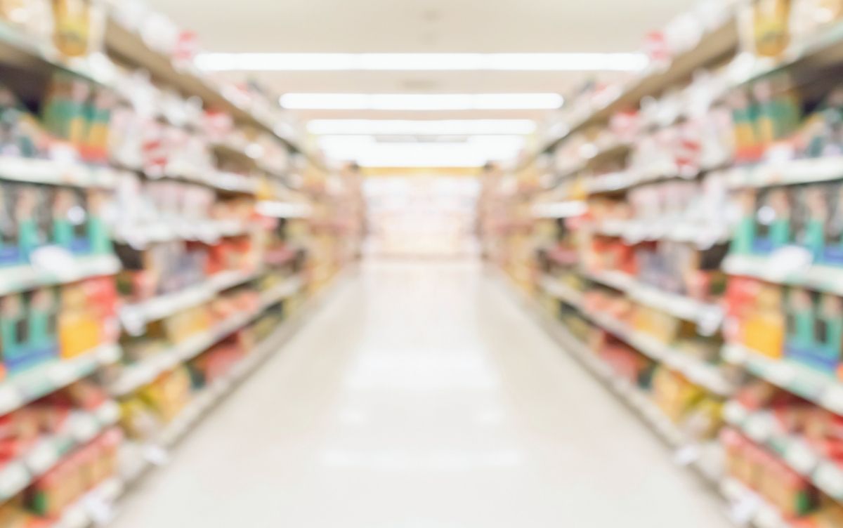 Blurred view of a brightly lit supermarket aisle with shelves stocked on both sides, suggesting a clean and well-maintained retail environment.