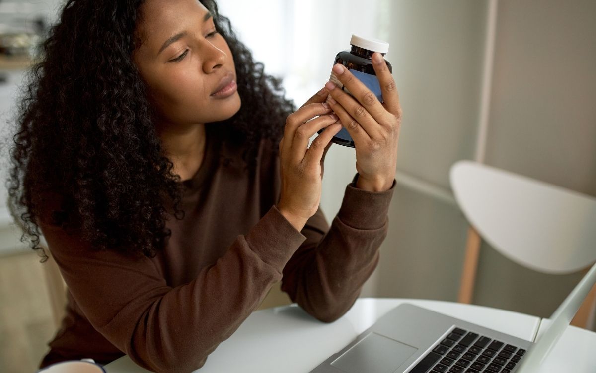 A person examines a medication bottle closely while sitting at a desk with a laptop.