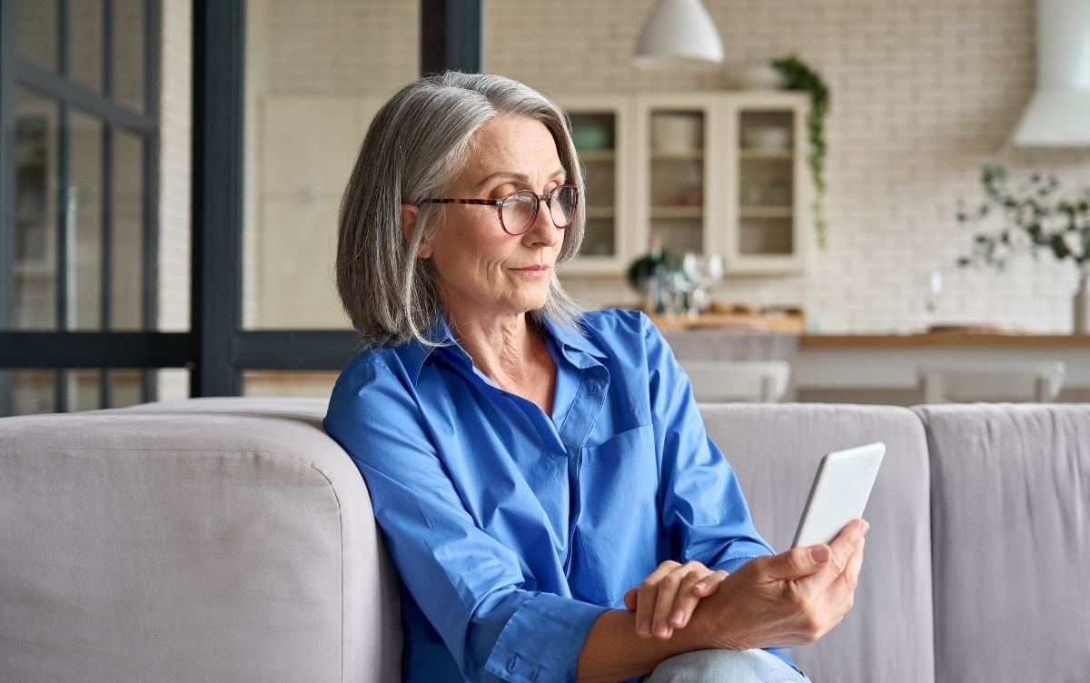 Person with gray hair sitting on a couch, wearing a blue shirt and holding a smartphone, with a modern kitchen featuring white cabinets and plants in the background.