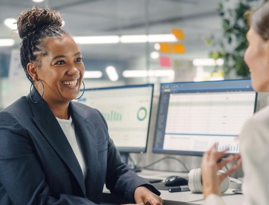 Two female colleagues have a conversation. One is smiling and the other has her back to the camera.