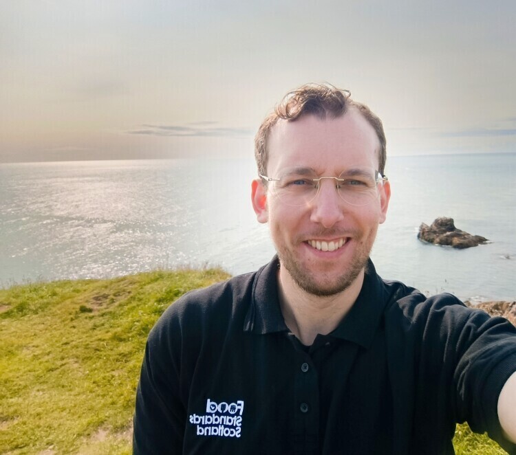 Ntooukan Achmetoglou. He's smiling looking at the camera and wearing a polo shirt with the Food Standards Scotland logo on it.